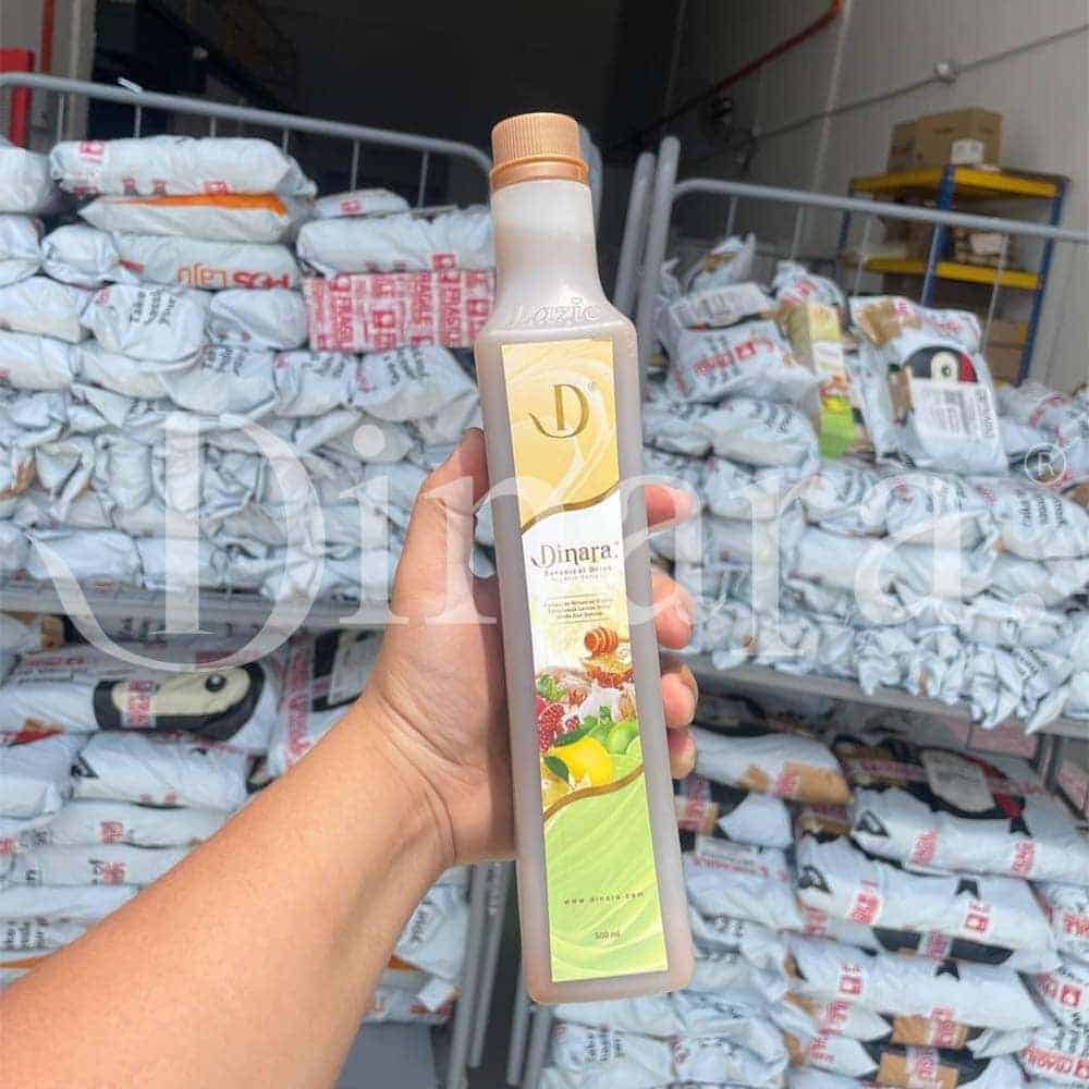 Bright hand holding Dinara honey and herbal syrup bottle in warehouse with stored bags.